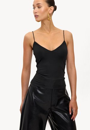 Femme portant un haut noir à fines bretelles et un pantalon en cuir noir brillant, regardant vers sa gauche, avec des boucles d'oreilles dorées en forme de créoles et les cheveux attachés en arrière.