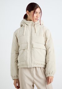 Bežová kordurová péřová bunda s kapucí, má přední zip, dvě náprsní kapsy, elastické manžety a zřasený lem.