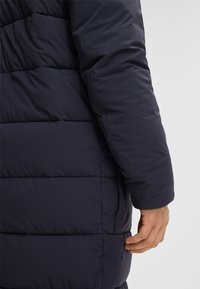Eine Person trägt eine marineblaue, gesteppte Jacke und zeigt die Seitenansicht des Arms und der Hand in der Nähe der Seitentasche der Jacke.