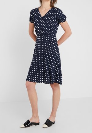 Kobieta w granatowej sukience do kolan w białe groszki, z krótkimi bufkami na rękawach, ubrana w czarno-białe wsuwane buty.