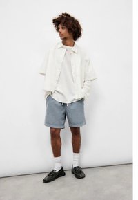 Ελαφρύ, εκρού πουκάμισο με κοντά μανίκια πάνω από λευκή υφασμάτινη μπλούζα, συνδυασμένο με μπλε σορτς. Μαύρα παπούτσια και λευκές κάλτσες ολοκληρώνουν την εμφάνιση.