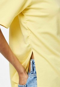 T-shirt di cotone gialla con maniche corte, fessure laterali e una vestibilità rilassata, parzialmente infilata in pantaloni corti di denim azzurro.