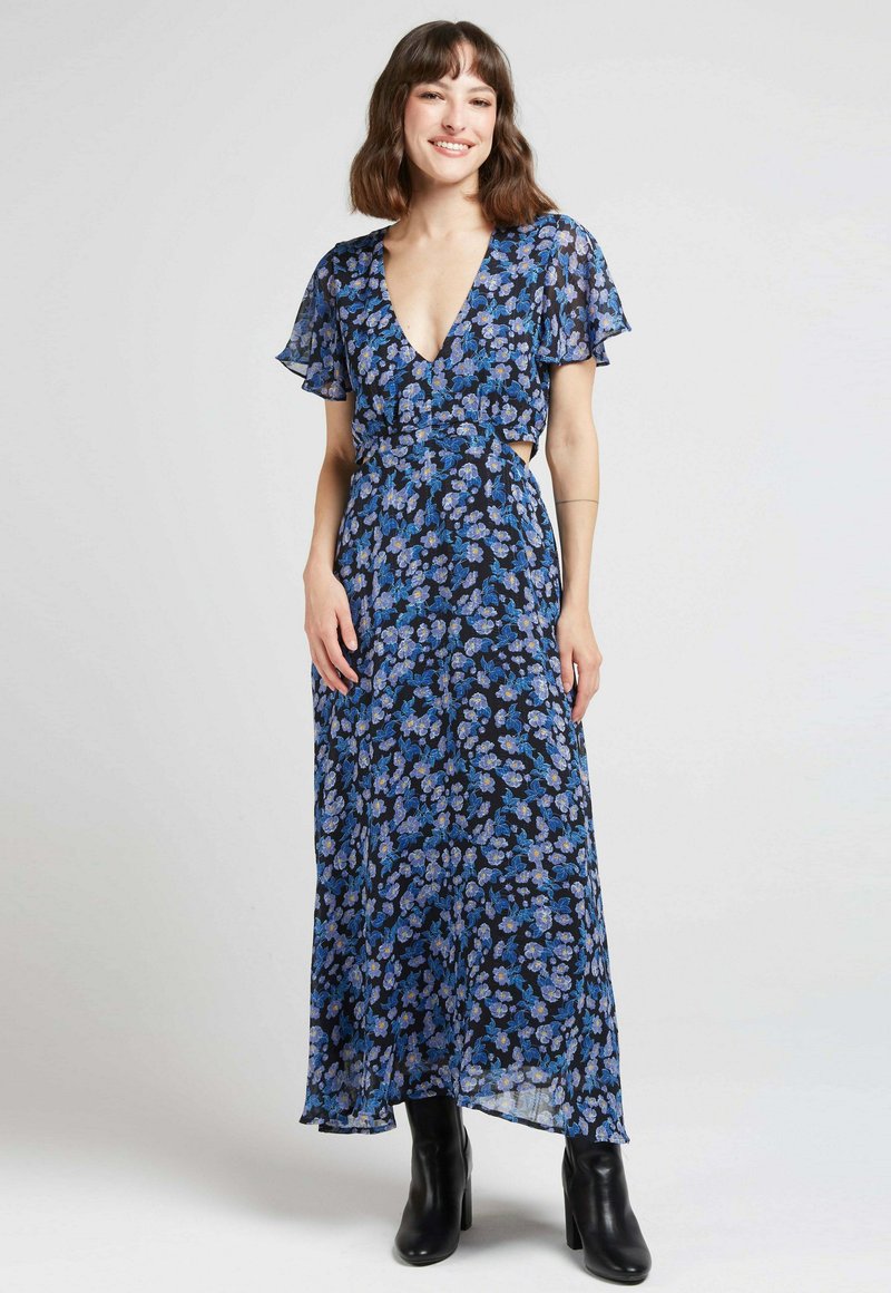 Robe fluide fleurie bleu marine avec des motifs bleus et jaunes, décolleté en V, manches courtes volantées et jupe fluide, associée à des bottes à talons noires.