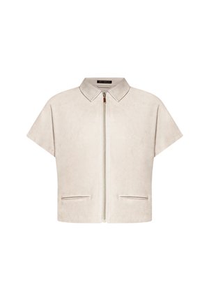 Kobieta ubrana w beżową kurtkę na krótki rękaw z zamkiem błyskawicznym na czarny top, beżowe szerokie spodnie z bocznymi kieszeniami oraz beżowe sandały na obcasie, stojąca z ręką w kieszeni.