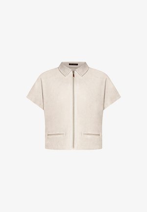 Kobieta ubrana w beżową kurtkę na krótki rękaw z zamkiem błyskawicznym na czarny top, beżowe szerokie spodnie z bocznymi kieszeniami oraz beżowe sandały na obcasie, stojąca z ręką w kieszeni.