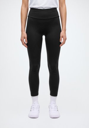 Un modèle porte un legging noir taille haute, un haut blanc, des chaussettes et des baskets, et se tient devant un fond uni clair.