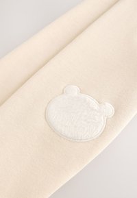 Tissu beige avec un patch blanc et duveteux en forme de visage d'ours cousu près du bord central.