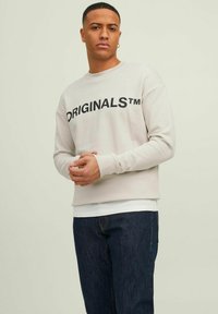 Light gray sweatshirt with "ORIGINALS™" printed in bold black letters. Soft material, crew neck, and ribbed cuffs. Paired with dark denim jeans.