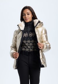 Skinnende guld puffjakke med pelsforet hætte, sort lynlåstop med et geometrisk mønster, parret med sorte tætsiddende bukser.