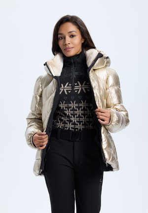 Schillernde goldene Pufferjacke mit fellgefütterter Kapuze, schwarzes Reißverschluss-Oberteil mit geometrischem Muster, kombiniert mit schwarzen, engen Hosen.