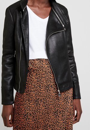 Personne portant un t-shirt blanc à col en V, une veste en cuir noire avec des fermetures éclair argentées et une jupe noire avec des taches orange irrégulières.
