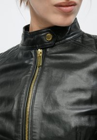 Μαύρο δερμάτινο μπουφάν με φερμουάρ, ψηλό γιακά και χρυσές μεταλλικές λεπτομέρειες, με καπιτονέ διακόσμηση στους ώμους.