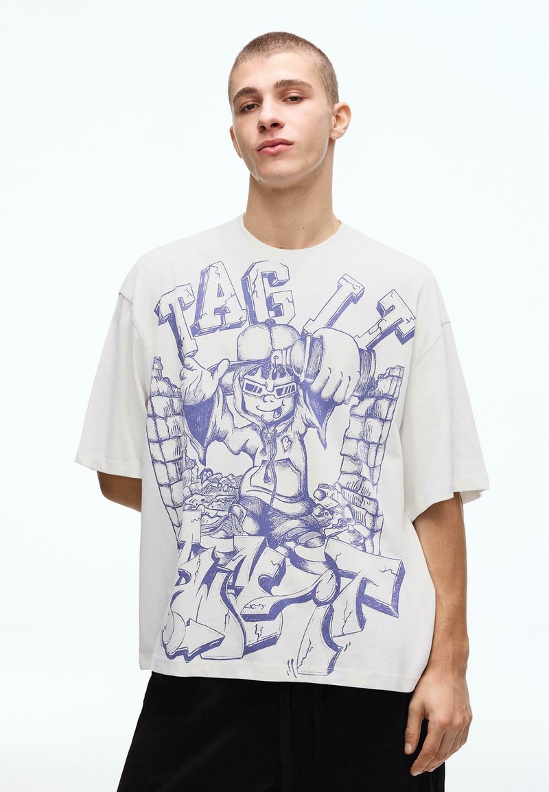 Junger Mann trägt ein weißes, übergroßes T-Shirt mit einer blauen Graffiti-ähnlichen Cartoonfigur und dem fett gedruckten Text "TAG".