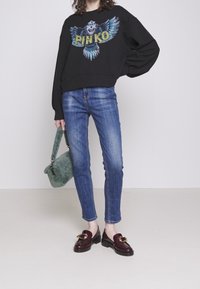 Felpa nera con grafica di un gufo colorato, jeans blu aderenti, mocassini bordeaux scuri con hardware dorato e una borsa verde pelosa.