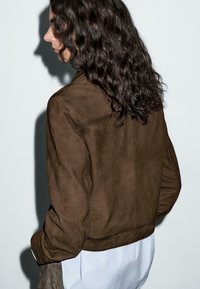 Brown suede jacket with a minimalist design, featuring a cropped cut and side pockets, worn over a white shirt. Textured finish.