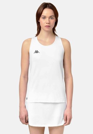 Jeune femme portant un débardeur de sport blanc et une jupe assortie, debout devant un fond clair uni.