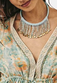 Collar de cuentas con cuerdas azul claro y monedas doradas, colgando en un diseño de flecos. Llevado con una prenda con patrón floral y borde de encaje.
