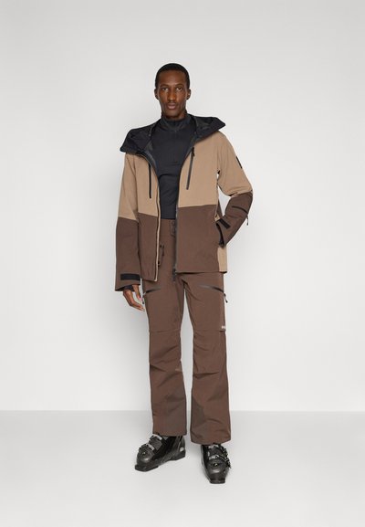 Brun skidjacka med en ljusbrun övre panel, svarta detaljer och matchande bruna byxor. Båda har blixtlås och en slitstark textur.