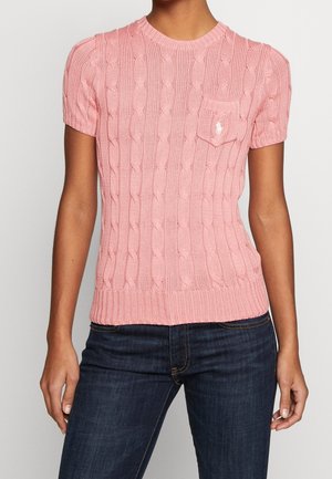 Femme portant un pull en maille torsadée rose ajusté à manches courtes avec une petite poche poitrine et un jean bleu foncé.
