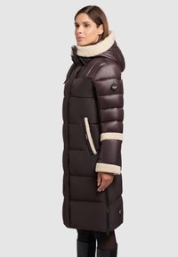 Giacca a vento lunga marrone con colletto e polsini in pelliccia sintetica color crema. Presenta dettagli con zip e tasche, e una texture leggermente lucida.