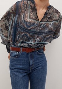 Blouse transparente à motifs dans des tons bleus, bruns et crème avec un décolleté en V, associée à un jean bleu taille haute et une ceinture marron.