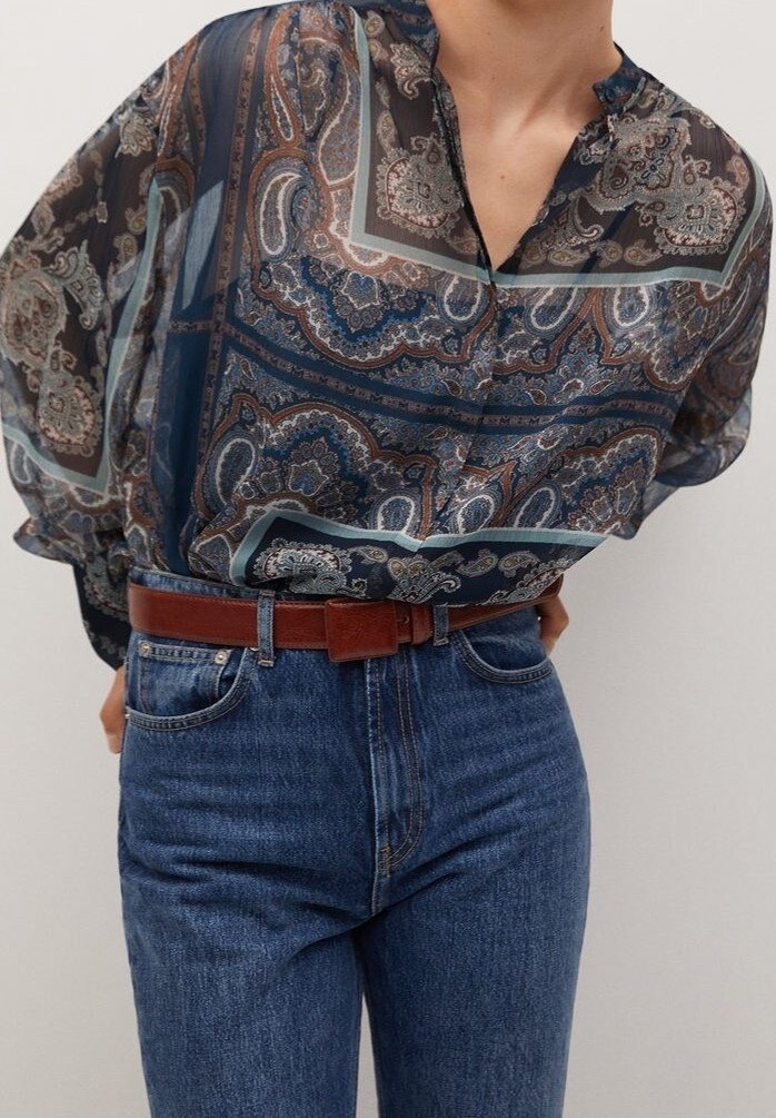 Blouse transparente à motifs dans des tons bleus, bruns et crème avec un décolleté en V, associée à un jean bleu taille haute et une ceinture marron.