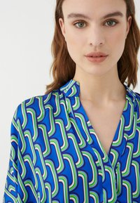 Blusa azul con un patrón geométrico en verde, rosa y blanco. Presenta un diseño sin cuello y una textura de tela suave.