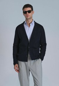 Blazer en tricot bleu marine avec deux poches avant, fermeture à boutons, superposé sur une chemise à carreaux bleu clair et un pantalon gris plissé. Lunettes de soleil noires.