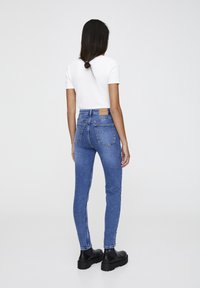Jeans ajustados de denim azul de cintura alta con un acabado texturizado, bolsillos traseros y una etiqueta de cuero marrón en la cinturilla. Botas negras gruesas.