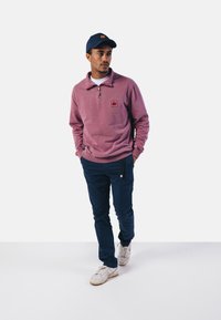 Pull en tissu rose doux avec zip quart, petite patch logo sur la poitrine ; associé à un pantalon marine et des baskets blanches. Casquette marine sur la tête.
