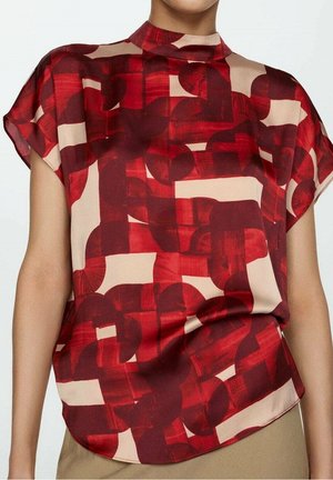 Kobieta ubrana w bluzkę z krótkim rękawem o czerwono-beżowym geometrycznym wzorze, z wysokim, okrągłym dekoltem i luźnym krojem.