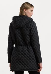 Veste matelassée noire avec une capuche, dotée d'une taille à cordon de serrage et d'un motif en losanges. Tissu texturé avec des manches longues et une coupe mi-longue.