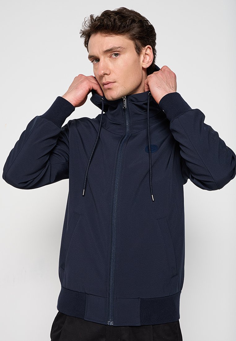 Giovane che aggiusta il cappuccio su una giacca blu navy con zip e tasche frontali, in piedi contro uno sfondo chiaro e semplice.