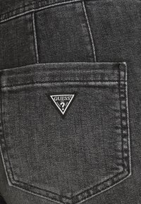 Bolsillo de mezclilla negra con un parche triangular del logo "GUESS" que presenta detalles de costura, tela texturizada y un acabado de lavado oscuro.