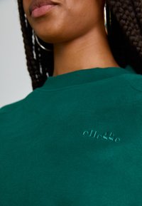 Sudadera verde de cuello redondo hecha de tejido suave, con un logo bordado tonal en el pecho y un cuello acanalado.
