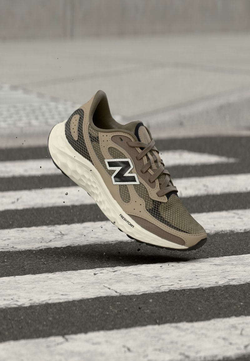Καφέ και μαύρο παπούτσι τρεξίματος New Balance με λευκή σόλα, αιωρούμενο πάνω από διάβαση πεζών σε οδόστρωμα από άσφαλτο.