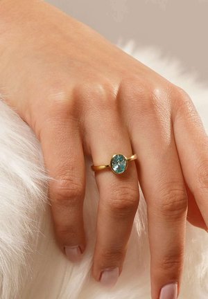 Hand mit goldring mit ovalem blauem Edelstein am Ringfinger, ruhend auf weichem weißem Fellhintergrund.