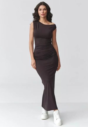 Kobieta o długich ciemnych włosach, ubrana w dopasowaną, asymetryczną, bezrękawnikową czarną suknię maxi oraz białe sneakersy na platformie, stojąca na jednolitym tle.