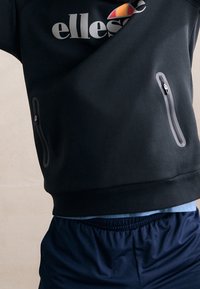 Sweat-shirt noir avec fermetures éclair grises de chaque côté et logo sur la poitrine, associé à un short bleu marine avec une taille élastique.