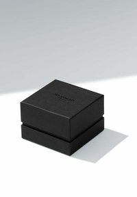 Svart texturerad fyrkantig låda med lock, med präglad branding på toppen. Lådan har en minimalistisk design och en slät yta.