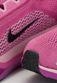 Nike atletska cipela s gorenjim dijelom od ružičaste mreže, crnim swoosh logotipom, teksturiranim detaljima i ljubičastom petom. Ima podstavljeni đon za podršku.