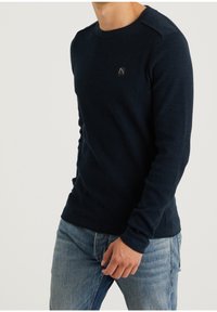 Granatowy sweter z teksturowanym splotem, okrągłym dekoltem i detalem na ramieniu. Posiada małe czarne logo na klatce piersiowej i dopasowane rękawy.