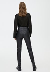 Pantalons en faux cuir noirs taille haute avec un design élégant, deux poches arrière et des détails de couture subtils ; associés à un haut transparent noir à manches longues.