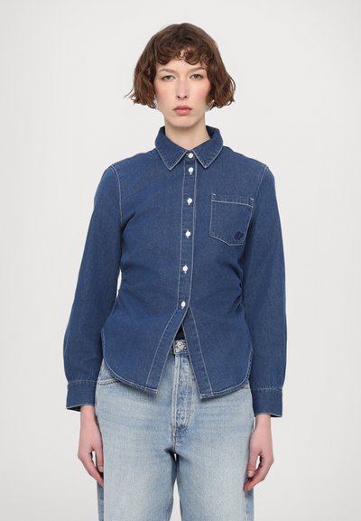 Camisa de mezclilla en azul oscuro, con cuello abotonado, bolsillo delantero y costuras blancas. Tiene un corte con dobladillo curvado y mangas largas.
