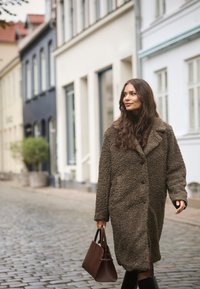 Palton mare, texturat, de culoare maro, cu nasturi și guler. Poartă o geantă de mână maro închis și merge pe o stradă cu piatră cubică.