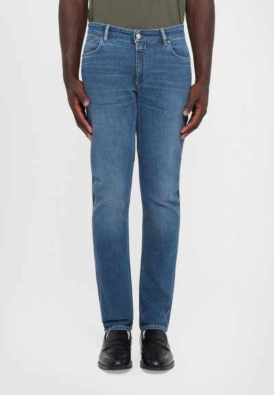 Homme portant un jean bleu slim et des mocassins en cuir noirs, se tenant contre un fond blanc uni.