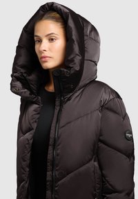 Puffer jacket in dark brown, featuring a large, folded hood, zip front, and a circular logo patch on the left arm. Smooth, quilted texture.