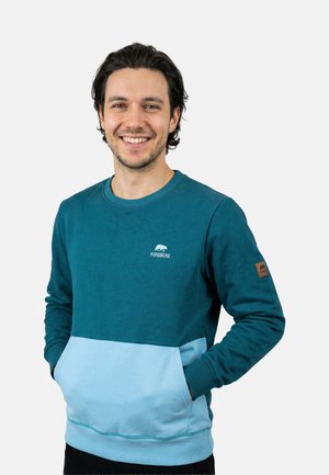 Smiling man with dark hair wearing a teal and light blue Forsberg sweatshirt with hands in front pocket, standing against white background.