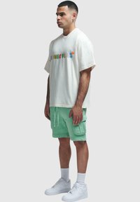 Cremfarbener, oversized T-Shirt mit mehrfarbigem Grafiktet, kombiniert mit mintgrünen Cargo-Shorts und weißen Sneakern. Einfaches und lässiges Design.