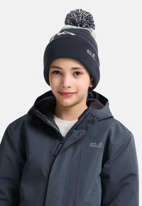 Dunkelblauer gestrickter Beanie mit weißen Mustern, oben mit einem Pompon; getragen mit einer dunklen Jacke mit Reißverschluss und Logo.